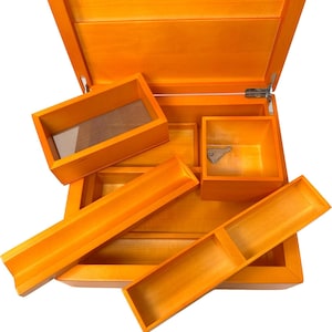 Può includere: Una scatola di legno aperta, tinta di arancione, con diversi vassoi e scomparti rimovibili. La scatola ha un coperchio incernierato e una chiusura in metallo. L'interno presenta un pezzo di metallo a forma di chiave. I vassoi sono di varie dimensioni e forme.