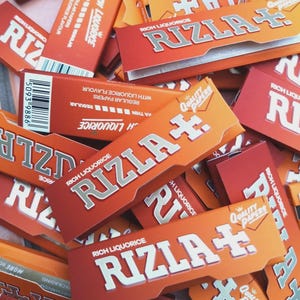 Puede incluir: Un surtido de paquetes de papel de liar Rizla de color naranja y rojo. Los paquetes presentan el nombre de la marca en letras plateadas grandes, con la frase "Rich Liquorice" encima. Algunos paquetes tienen el texto "Quality Papers" y "Regular Papers".