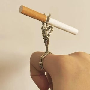 May include: A silver-colored ring shaped like a skeleton hand holding a lit cigarette. The ring is worn on a finger, with the cigarette held upright. The cigarette has a brown filter and white paper.