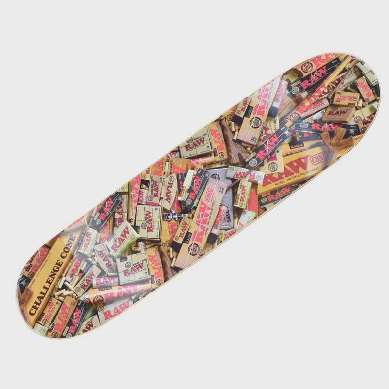 May include: A skateboard deck covered in a collage of RAW rolling paper boxes and related products. The design features various sizes and styles of packaging, with text like "CHALLENGE CONE" and "RAW" visible. The deck has a light wood color.