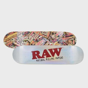 May include: Two skateboards. The top one has a collage of rolling paper boxes. The bottom one is white with the word "RAW" in red and the text "NATURAL ROLLING PAPERS" below it.