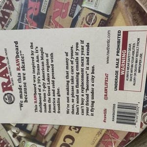 May include: A round skateboard deck with a design featuring the RAW logo and various rolling paper packaging. The deck has a paper label with text about the product's origin and a warning about underage sales.