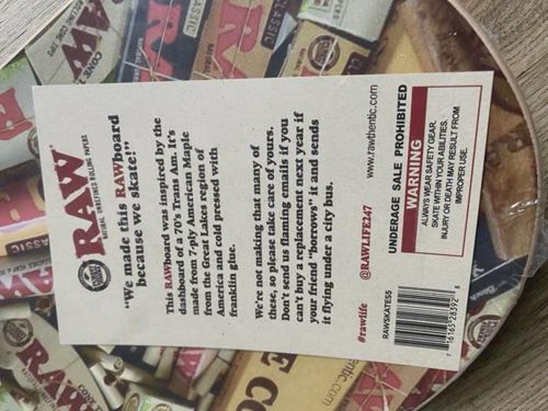 May include: A round skateboard deck with a design featuring the RAW logo and various rolling paper packaging. The deck has a paper label with text about the product's origin and a warning about underage sales.