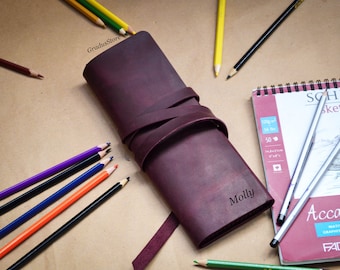 May include: A burgundy leather pencil case with a strap closure and the name "Molly" engraved on the front. The case is lying on a brown surface with colored pencils and a sketchbook.