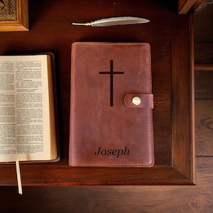 May include: A brown leather journal with a black cross and the name "Joseph" printed on the cover. The journal has a snap closure and is on a wooden surface next to an open book and a feather.