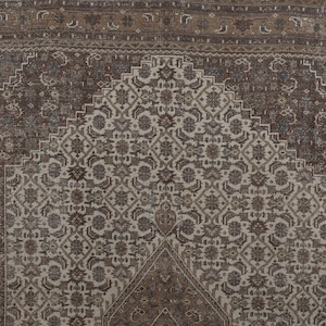 May include: A detailed, close-up view of a vintage rug. The rug features a central design in cream and brown tones, with intricate floral and geometric patterns. The border is a darker brown, framing the central design. The rug's texture and design suggest it is a traditional, handmade piece.