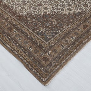 May include: A corner view of a rectangular, vintage rug with a detailed, floral pattern. The rug features a border in shades of brown and beige, with a central field in similar tones. The design includes geometric and floral motifs.
