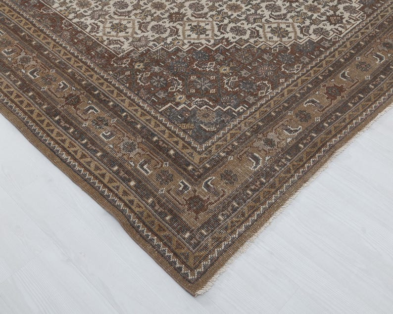 May include: A corner view of a rectangular, vintage rug with a detailed, floral pattern. The rug features a border in shades of brown and beige, with a central field in similar tones. The design includes geometric and floral motifs.