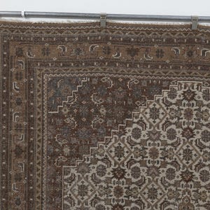 May include: A section of a brown and beige vintage rug with an intricate geometric pattern. The rug has a detailed border and a central design featuring floral and geometric shapes. The rug is hanging from a metal bar with clips.