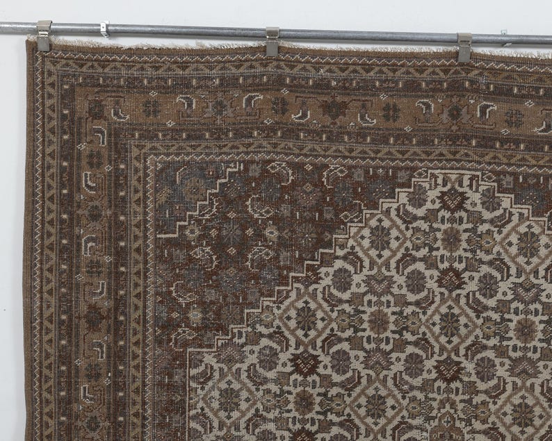 May include: A section of a brown and beige vintage rug with an intricate geometric pattern. The rug has a detailed border and a central design featuring floral and geometric shapes. The rug is hanging from a metal bar with clips.