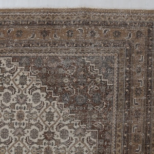 May include: Close-up of a section of a vintage rug with a detailed, ornate design. The rug features a central pattern in cream and brown tones, surrounded by a border in shades of brown and tan. The intricate patterns and color palette suggest a traditional style.