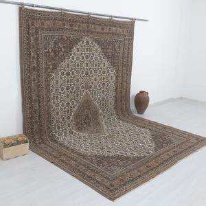 May include: A large, rectangular, brown and beige rug with an intricate floral pattern. The rug is hanging from a metal rod, with a decorative border and a central design. A small wooden chest and a brown vase are in the background.