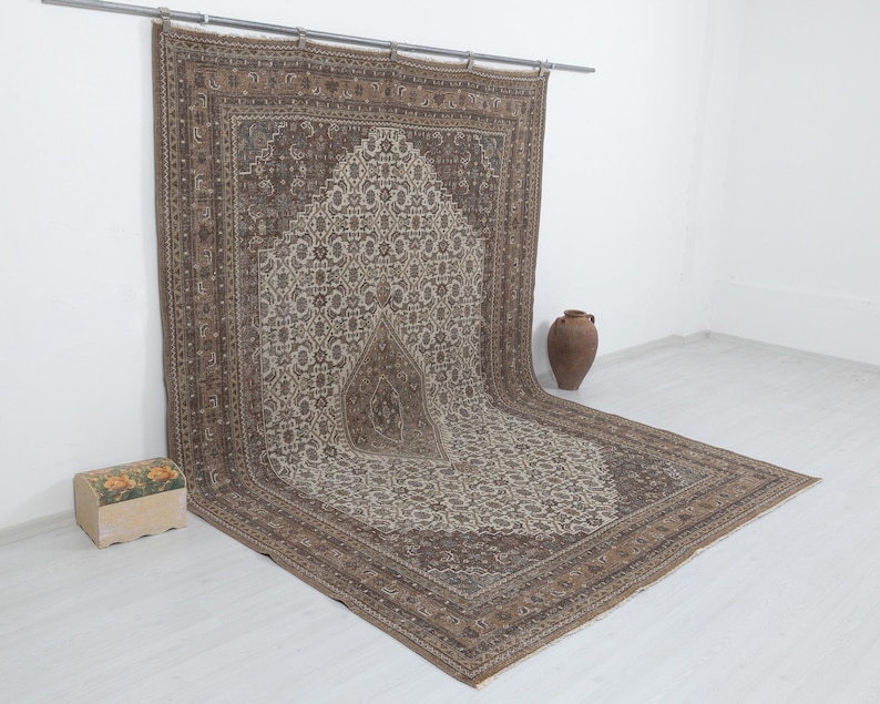 May include: A large, rectangular, brown and beige rug with an intricate floral pattern. The rug is hanging from a metal rod, with a decorative border and a central design. A small wooden chest and a brown vase are in the background.