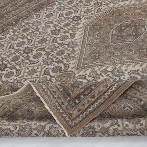 May include: A vintage-style area rug with a detailed floral and geometric pattern. The rug features a palette of brown, beige, and cream tones, with a folded corner revealing the fringed edge. The design includes a central medallion and border.