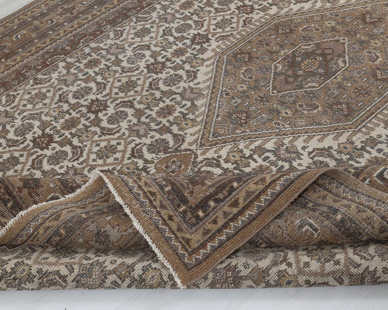 May include: A vintage-style area rug with a detailed floral and geometric pattern. The rug features a palette of brown, beige, and cream tones, with a folded corner revealing the fringed edge. The design includes a central medallion and border.