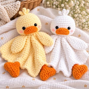Può includere: Due doudou all'uncinetto a forma di anatra. Un'anatra è giallo pallido e l'altra è bianca. Entrambe hanno becchi arancioni, piedi arancioni, occhi di sicurezza neri e piccole macchie rosa sulle guance. I corpi sono in filato morbido e testurizzato.