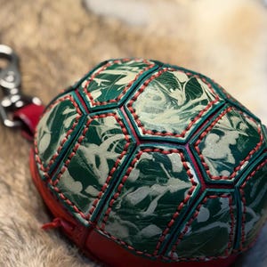 May include: A turtle-shaped leather pouch featuring a shell constructed from hexagonal panels with a marbled green and cream pattern. The panels are joined by red stitching. A red leather strap with a silver metal swivel clasp is attached to the side.