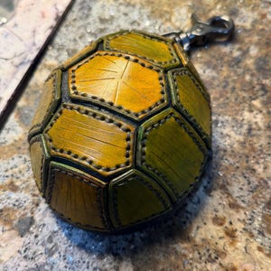 May include: A rounded leather pouch featuring a segmented, turtle-shell-inspired design in shades of yellow and green. The surface has visible stitching along the edges of each panel and includes a metal swivel lobster clasp attached to the top.