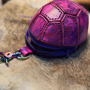 May include: A small, dome-shaped pouch with a marbled purple and pink finish, constructed from stitched leather panels resembling a turtle shell. The pouch features a dark purple zipper closure and an iridescent, multi-colored metal swivel clasp.