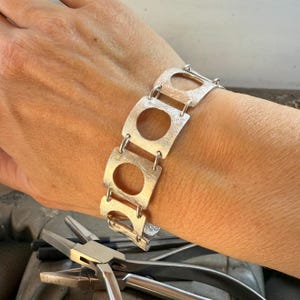 Könnte beinhalten: Ein silbernes Armband mit geometrischem Design. Das Armband besteht aus miteinander verbundenen rechteckigen Formen mit kreisförmigen Ausschnitten.