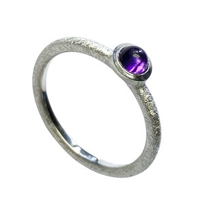 Könnte beinhalten: Ein silberner Ring mit einer strukturierten Band und einem runden, violetten Amethyst-Edelstein.