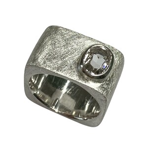 Könnte beinhalten: Ein silberner Ring mit gebürsteter Metallstruktur und einem großen, oval geschliffenen, klaren Edelstein. Der Ring hat ein breites Band und ein modernes, geometrisches Design. Der Edelstein ist in einer Zargenfassung gefasst.