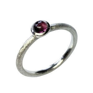 Könnte beinhalten: Ein silberner Ring mit einem kleinen, runden, violetten Edelstein, der in einer Zargenfassung gefasst ist.