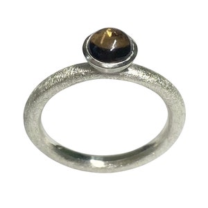 Könnte beinhalten: Silberring mit strukturiertem Band und einem runden, dunkelbraunen Edelstein. Der Edelstein ist in einer silbernen Lünette gefasst, wodurch ein schlichtes, aber elegantes Design entsteht. Ein klassisches Schmuckstück.