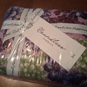 May include: A folded bundle of fabric featuring a floral pattern in shades of purple, pink, and green. The fabric is wrapped in white ribbon printed with the text Cut out for happiness and includes a white tag labeled Boundless FABRICS.