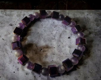 May include: A purple and iridescent square-shaped beaded bracelet with a silver-toned metal clasp.