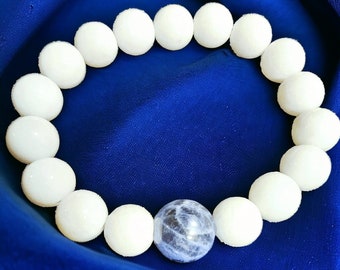 May include: A white beaded bracelet with a blue gemstone charm. The bracelet is made of round, white beads and has a single, round, blue gemstone charm in the center.