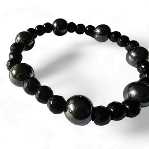 Op de afbeelding: Een zwarte kralen armband met afwisselende grote en kleine kralen. De grote kralen zijn rond en hebben een glanzende, metallic afwerking. De kleine kralen zijn gefacetteerd en hebben een matte afwerking.