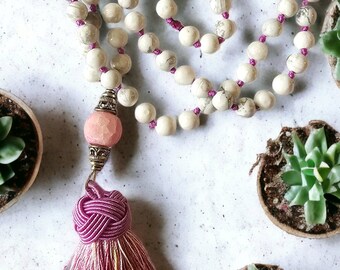 Op de afbeelding: Een witte en grijze stenen mala-ketting met een roze kwast. De ketting is gemaakt van ronde kralen en is geregen aan een roze koord. De kwast is gemaakt van roze en gele draden.
