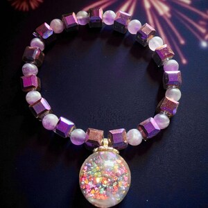 Könnte beinhalten: Ein Armband mit irisierenden lila Würfelperlen und hellvioletten runden Perlen. Ein klarer, runder Anhänger, gefüllt mit bunten Glitzer, hängt an dem Armband. Das Armband befindet sich vor einem dunklen Hintergrund mit einem Feuerwerksdesign.