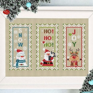 May include: Three cross-stitch patterns featuring a snowman, Santa Claus, and a reindeer. The patterns are framed in white and have a beige background. The snowman pattern says "SNOW", the Santa Claus pattern says "HO! HO! HO!" and the reindeer pattern says "JOY".