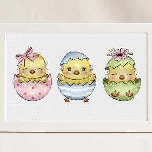 Può includere: Un ricamo a punto croce incorniciato con tre pulcini gialli che escono da uova decorate. Le uova sono rosa a pois bianchi, blu a strisce e verdi con fiori. Ogni pulcino ha gli occhi chiusi e un piccolo becco.