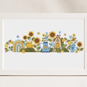 Könnte beinhalten: Kreuzstichmuster mit einer fantasievollen Gartenszene mit Sonnenblumen, einem Frosch, einer Gießkanne, einem kleinen Haus und einem Regenbogen. Das Muster ist in einem weißen Rahmen.