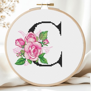 Op de afbeelding: Een borduurring met kruissteek met de zwarte letter "C", versierd met roze rozen en groene bladeren. Het bloemmotief staat tegen een witte achtergrond, omlijst door een houten ring.
