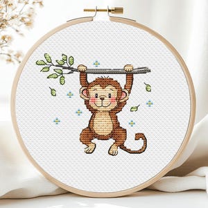 May include: A cross-stitch embroidery hoop featuring a cartoon monkey hanging from a branch. The monkey is brown with pink cheeks, and the branch has green leaves. The background is white, and the hoop is made of wood.