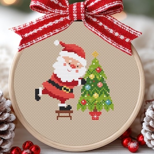Può includere: Un telaio da ricamo a punto croce con Babbo Natale che decora un albero di Natale. Babbo Natale indossa un abito e un cappello rossi, l'albero è verde con ornamenti colorati. Un fiocco a righe rosse e bianche adorna la parte superiore.