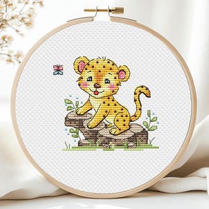 May include: A cross-stitch embroidery hoop featuring a cute cartoon leopard cub with yellow fur and brown spots. The cub sits on a rock with green grass and blue flowers. A small butterfly is in the upper left corner.
