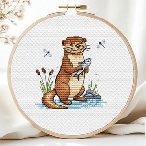 Könnte beinhalten: Ein Kreuzstich-Stickrahmen mit einem braunen Otter, der einen Fisch hält. Der Otter steht im Wasser mit Felsen und Schilf. Zwei blaue Libellen sind im Hintergrund. Die Stickerei befindet sich in einem Holzrahmen.