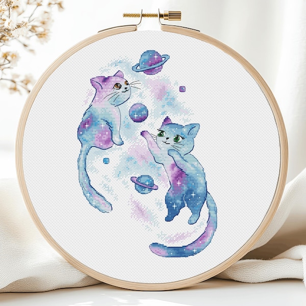 May include: Two blue and purple cats with galaxy patterns play with a ball and a planet in a white embroidery hoop. The background is a white fabric with a galaxy pattern.