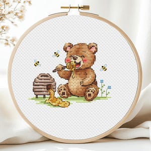 Könnte beinhalten: Kreuzstich-Stickerei in einem Holzrahmen zeigt einen braunen Teddybären, der Honig isst. Der Bär sitzt in der Nähe eines Bienenstocks, mit herumfliegenden Bienen. Das Design befindet sich auf weißem Stoff.