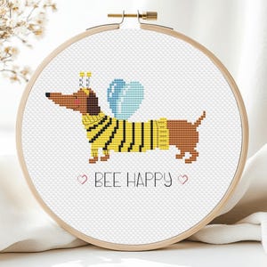 Könnte beinhalten: Kreuzstichmuster eines Dackels, der ein Bienenkostüm mit Flügeln trägt und den Text "BEE HAPPY" in rosa Herzen.
