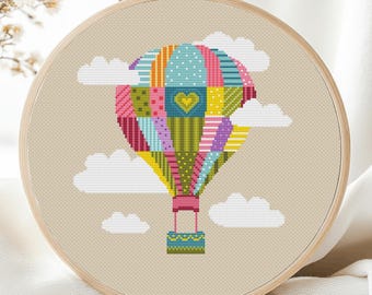 May include: A colorful cross-stitch of a hot air balloon with a heart design, set against a neutral background with white clouds. The balloon features a patchwork design in various colors, including pink, blue, yellow, and green, framed in a wooden embroidery hoop.
