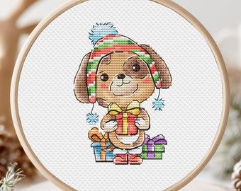 May include: Cross-stitch embroidery of a cartoon puppy wearing a striped hat and holding a wrapped gift. The puppy is surrounded by other wrapped gifts in blue, green, and purple. The embroidery is set in a wooden hoop.
