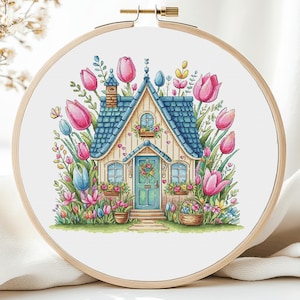 Może przedstawiać: Tamborek do haftu krzyżykowego z fantazyjną sceną wiejskiego domku. Domek ma niebieski dach, zielone drzwi i jest otoczony różowymi i niebieskimi tulipanami oraz innymi kwiatami. Haft jest umieszczony w drewnianym tamborku.