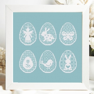 Puede incluir: Seis huevos de Pascua bordados blancos con diferentes diseños sobre un fondo azul claro. Los diseños incluyen una mariposa, un pájaro, un conejito, una flor, un ángel y un conejito con un corazón.