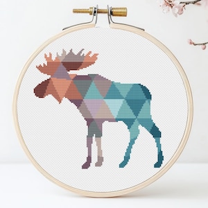 May include: A cross-stitch pattern of a moose with a geometric design in shades of blue, brown, and purple. The moose is standing on a white background.